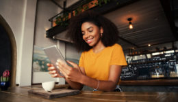 Low angle shot of beautiful African American female holding digital tablet sitting at funky tearoom with hot latte