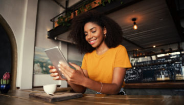 Low angle shot of beautiful African American female holding digital tablet sitting at funky tearoom with hot latte
