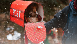 woman holds a red polka dot retro mailbox with Christmas gifts toys as well as a congratulatory letter from Santa on a background of fir trees.