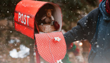 woman holds a red polka dot retro mailbox with Christmas gifts toys as well as a congratulatory letter from Santa on a background of fir trees.