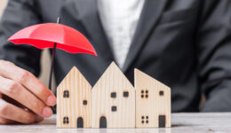 Businessman hand holding red Umbrella cover wooden Home model on table office. Property insurance and real estate concepts