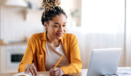 Focused cute stylish african american female student with afro dreadlocks, studying remotely from home, using a laptop, taking notes on notepad during online lesson, e-learning concept, smiling