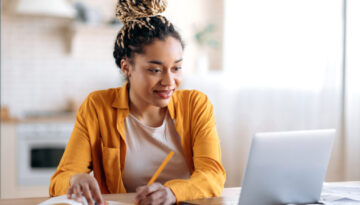 Focused cute stylish african american female student with afro dreadlocks, studying remotely from home, using a laptop, taking notes on notepad during online lesson, e-learning concept, smiling