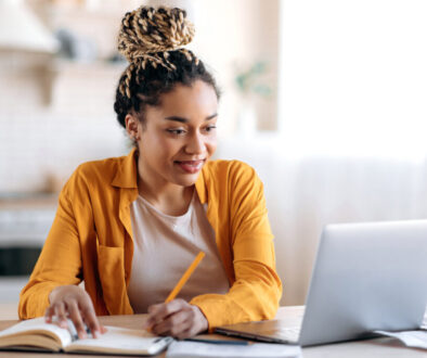 Focused cute stylish african american female student with afro dreadlocks, studying remotely from home, using a laptop, taking notes on notepad during online lesson, e-learning concept, smiling
