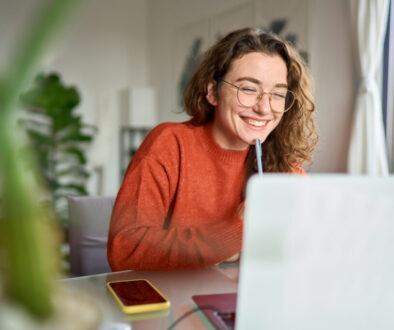 Young happy woman student using laptop watching webinar writing at home.