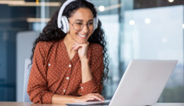 Young beautiful woman in headphones working with laptop inside office at workplace, joyful and satisfied business woman watching online webinar, educational video course, learning remotely