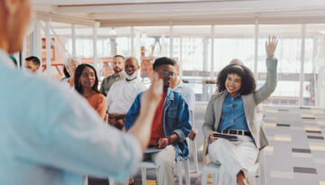 Speaker, crowd questions and happy woman with raised hand for presentation speech, team feedback or training workshop. Convention coach, conference meeting and business group answer at audience event