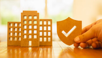 Agent holds insurance symbol next to a company building or condo building model, an idea about getting insurance in a condo or group insurance for company employees.